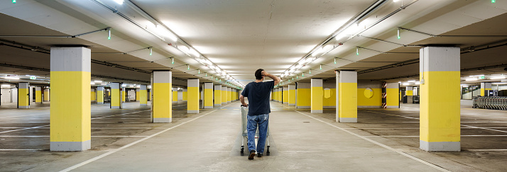Confused man walks with a shopping cart in an empty underground parking with yellow concrete columns and neon on the ceiling. Copy space