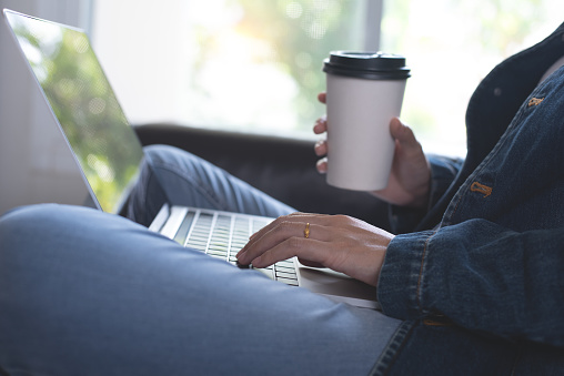 Young casual business woman holding cup of coffee, relax sitting on sofa and working, surfing internet on laptop computer at home, telecommuting, work from home concept