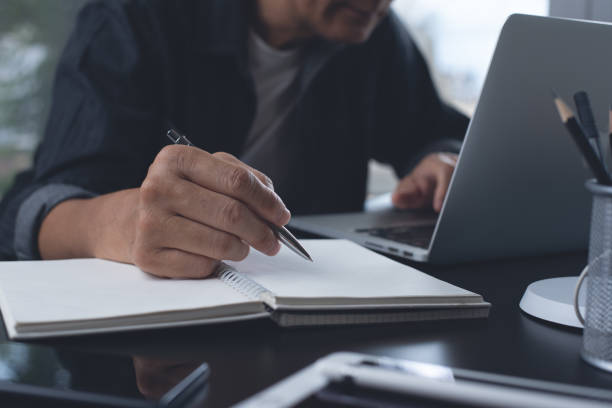 istock Casual business man with pen in hand, sitting at table writing on notebook, searching internet while working on laptop computer. Freelancer working at home. 1403090718