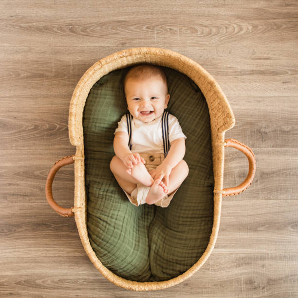 A 25-Week-Old Baby Boy Holding His Feet, Wearing a White Shirt With Navy Striped Suspenders & Khaki Shorts While Laying in a Cozy Green Cotton Blanket in a Seagrass Moses Basket Surrounded by Tropical House Plants for a Boho Tropical Vibe A 25-Week-Old Baby Boy Wearing a White Shirt With Navy Striped Suspenders & Khaki Shorts While Laying in a Cozy Cotton Blanket in a Seagrass Moses Basket Surrounded by Tropical House Plants for a Boho Tropical Vibe moses basket stock pictures, royalty-free photos & images