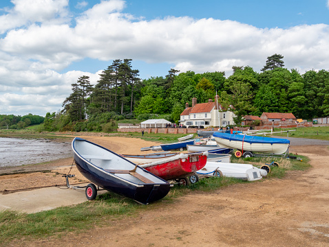 A row of small boats on the edge of the shore at Ramsholt on the Deben Peninsula in Suffolk, Eastern England. In the background is the Ramsholt Arms, a popular pub with locals and tourists.