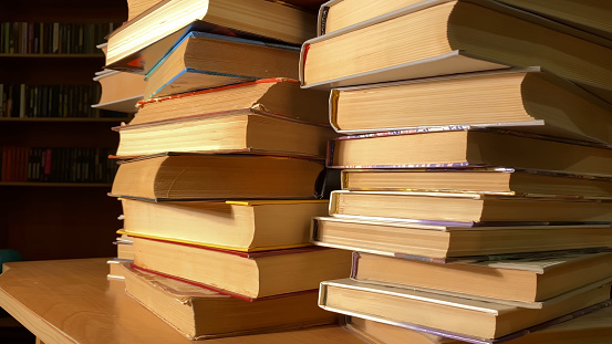 Stack of books on a wooden library shelf, the one on top open , multicolored book spines in a row in the background. Copy space on the left.