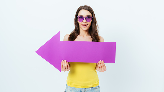 Portrait amazed young woman wearing purple glasses holds big arrow pointing to the side, white studio background