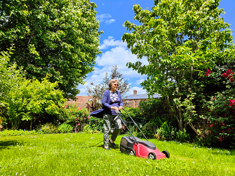 Active 80 year old Caucasian white female cutting the long grass with electric mower in her English country garden.