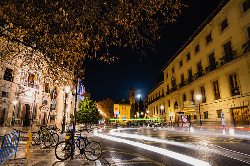 Long exposure photography of the Plaza de Santa Ana with traces of light from cars and some bicycles in the background, a church and the moon in the image at night