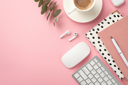 Business concept. Top view photo of workspace keyboard computer mouse stylish notepads pen cup of coffee on saucer earbuds and eucalyptus on isolated pastel pink background with copyspace