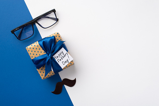 Father's Day concept. Top view photo of polka dot giftbox with ribbon bow and postcard glasses and mustache on bicolor blue and white background with copyspace