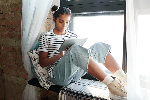 African girl sitting on windowsill and reading book online using digital tablet