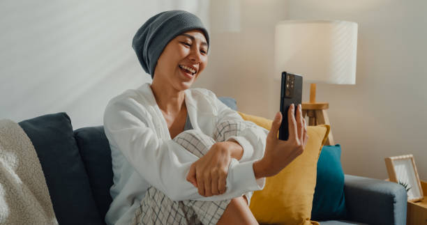 Young Asian woman sick with cancer using phone talk to friends sitting on sofa in living room at home. Young Asian woman sick with cancer using phone talk to friends sitting on sofa in living room at home. Telemedicine and Healthcare concept. cancer illness stock pictures, royalty-free photos & images