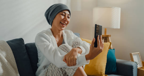 młoda azjatka chora na raka za pomocą rozmowy telefonicznej z przyjaciółmi siedzącymi na kanapie w salonie w domu. - patient women adult home interior zdjęcia i obrazy z banku zdjęć