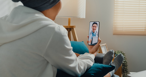 Young Asian woman sick with cancer using phone talk to doctor sitting on sofa in living room at home. Telemedicine and Healthcare concept.