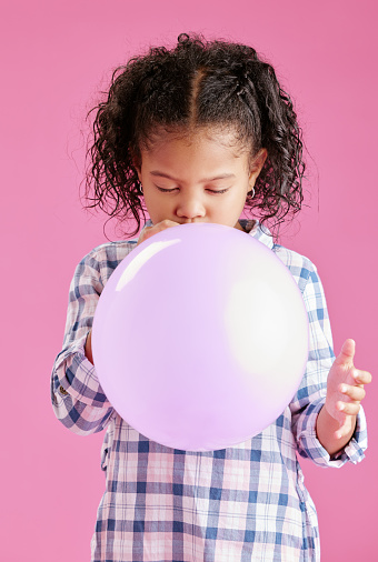 A pretty little mixed race girl with curly hair blowing a balloon against a pink copyspace background in a studio. African child looking content while having fun