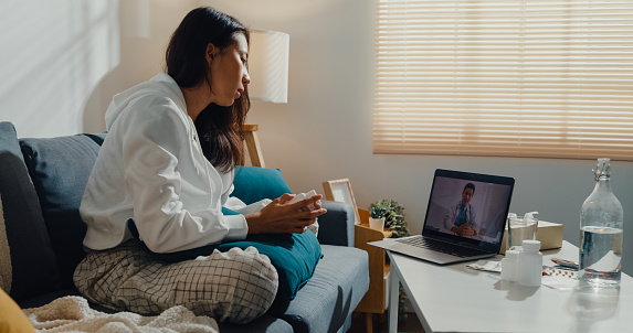 Sick young Asian woman using laptop talk to doctor sitting on sofa in living room at home. Telemedicine and Healthcare concept.