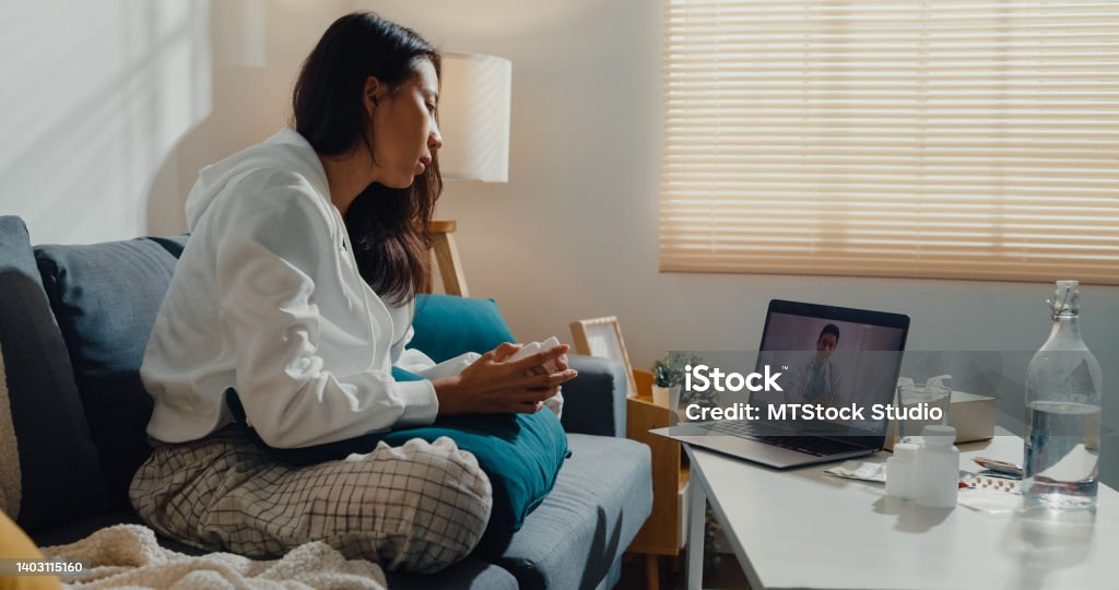 Sick young Asian woman using laptop talk to doctor sitting on sofa in living room at home. Sick young Asian woman using laptop talk to doctor sitting on sofa in living room at home. Telemedicine and Healthcare concept. Telemedicine Stock Photo