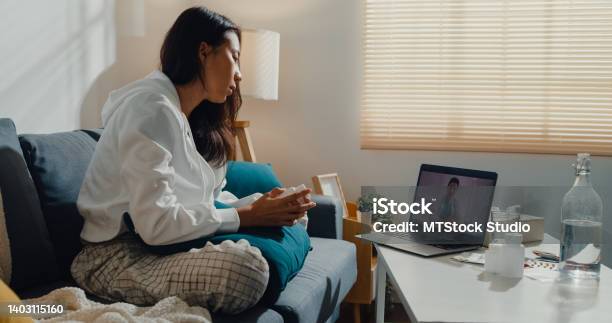 Sick Young Asian Woman Using Laptop Talk To Doctor Sitting On Sofa In Living Room At Home Stock Photo - Download Image Now