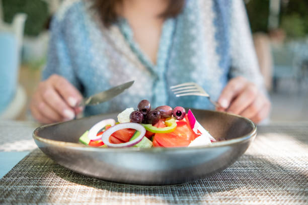 A woman with fork and knife eating vegan salad with tomatoes, cucumbers, slices of green pepper, red onion, tofu cheese and olives on top in her hands watching in camera. Woman defocused. Focus on salad.