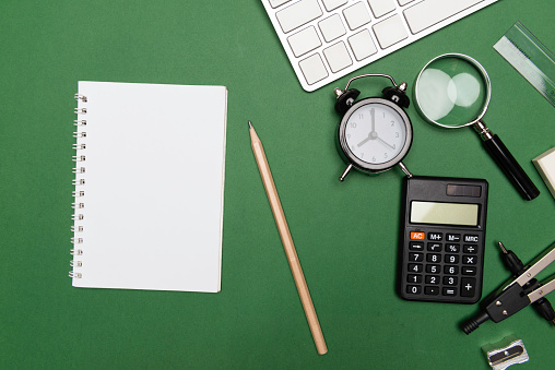 Directly above view of a green desk with office and school supplies like compasses, eraser, pen, magnifier, calculator,  sharpener, alarm clock, calculator,  notebook and a computer keyboard.Representing e-learning and distance working like home office.