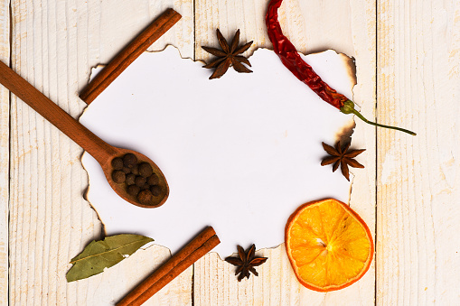 Composition of condiment on paper piece. Set of dry spices on beige wooden background. Food art concept. Wood spoon with black pepper balls, cinnamon, anise stars, dry orange slices and chili pepper