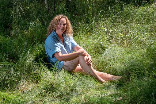 Forty year old woman sitting in the forest on the grass, laughing at the camera. She has wearing denim shirt.