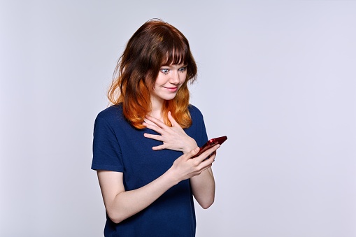 Beautiful smiling teenage female looking at smartphone screen, on light studio background. Positive red-haired girl with phone in her hands. Lifestyle, technology, communication, youth, young people