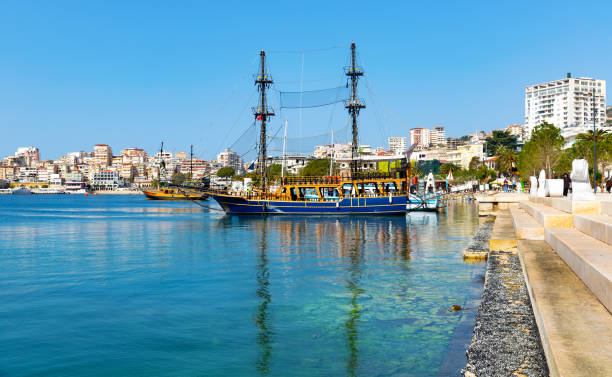 Scenic cityscape of Saranda on coast of gulf of Ionian Sea with vintage masted wooden sailing ships for sea trips moored near embankment on sunny day, Albania