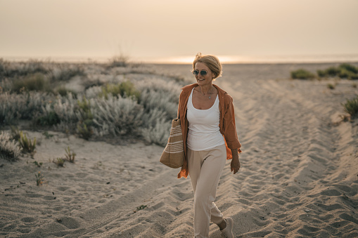 Mature woman carrying a bag is walking on the beach on a beautiful sunset.