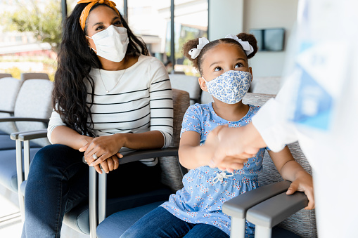In the clinic waiting room, the mid adult mother watches as her elementary age daughter shakes hands with the unrecognizable doctor.  Both mother and daughter wear protective masks.