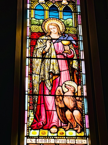 Vertical photo of the beautiful colourful stained glass window depicting Saint John the Evangelist, holding a golden goblet containing a purple dragon, with the Heraldic Eagle of St John at his feet, as seen from inside the heritage listed Saints Mary and Joseph Catholic Cathedral, Armidale, New England high country, northern NSW.