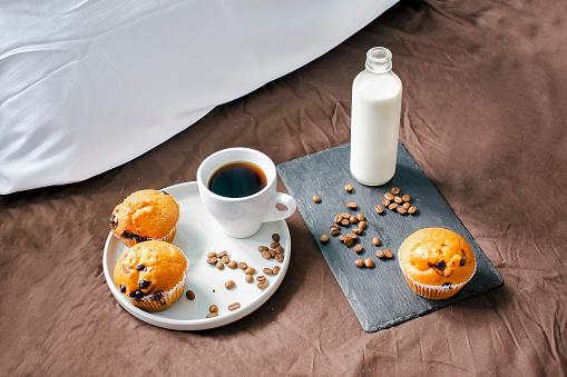 Cup of coffee, espresso and milk in bottle on the bed with muffins on plate, dessert for breakfast in berroom. sweet cozy home concept.