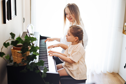 Mother teaching daughter to play piano. The girl is aged 6. Mother and girls are sitting by the old piano in a sunny room.