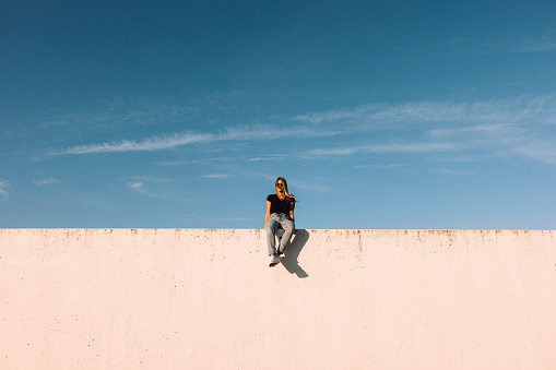 Young woman sitting on modern concrete wall with Sunglasses on. Simplicity Modern Urban Style. Shot from below against the sky. Urban Lifestyle Youth Portrait.
