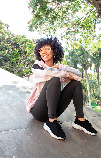 beautiful afro teen girl sitting in sportswear in park on sunny day and trees behind