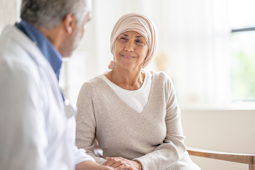 A female Cancer patient sits in the comfort of her own home as she meets with her Oncologist.  She is dressed comfortably in a sweater and wearing a headscarf to keep her warm.  Her male doctor is seated across form her in a white lab coat.