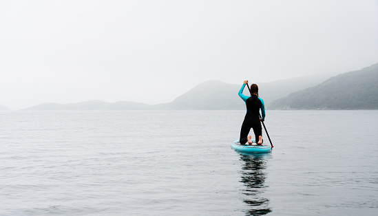 Back view active sportswoman in diving suit floating on stand up paddle at fog sea scenery