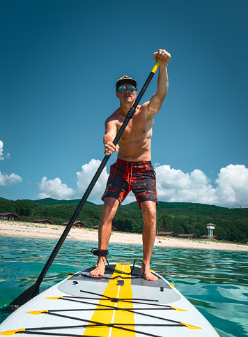 A tanned man stands on a yellow paddle board and paddles. Active sport and recreation on a sunny day on the beach in the turquoise sea water.
