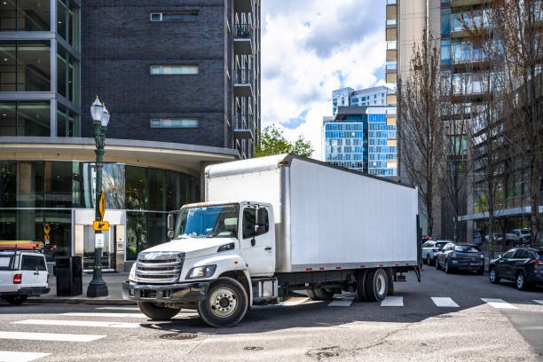 Big rig day cab white semi truck with long box trailer making local commercial delivery at urban city with multilevel residential apartments buildings turning on the city street with crossroad stock photo