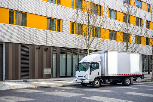 Small rig over cab white semi truck with long box trailer making local commercial delivery at urban city with multilevel residential apartments buildings standing on the street for upload cargo