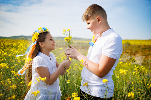 Kind teenage brother carefully hugs cheerful laughing sister with Ukrainian flower wreath with ribbons and smelling neat bouquet of flowers in hands, in yellow rapeseed blooming field under clear sky