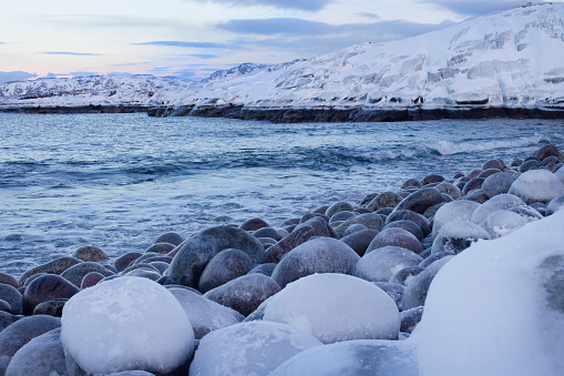 Winter seascape, beautiful sky and cold Barents Sea. Snow-covered rocky shore with large pebbles. Sea and blue sky. In the Far North. Russia, Teriberka, Kola Peninsula.