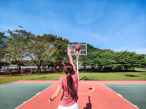 Rear view of woman practicing free throw and aiming at basketball hoop in outdoor court near residential area