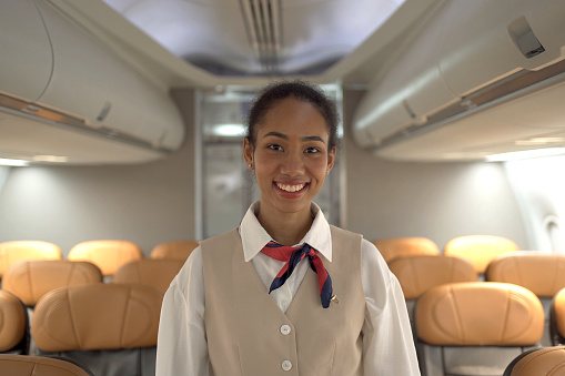 Beautiful female airline cabin crew looking and smiling to camera in the cabin. Confident and happy female flight attendee welcoming the passenger onboard.