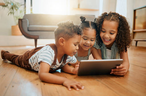 Three happy mixed race siblings relaxing on the lounge floor together while using a digital tablet. Three kids playing games on a digital device at home