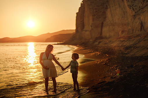 Happy single pregnant mother and her small boy holding hands and talking during summer sunset on the beach. Copy space.