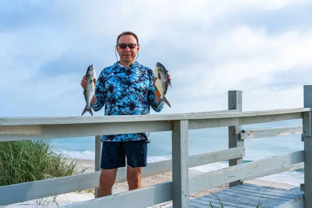 A white caucasian man 60 years old with a pompano and a bluefish in his hands stands on a wooden staircase against the backdrop of the Atlantic Ocean, having returned from fishing. Melbourne Beach, Florida