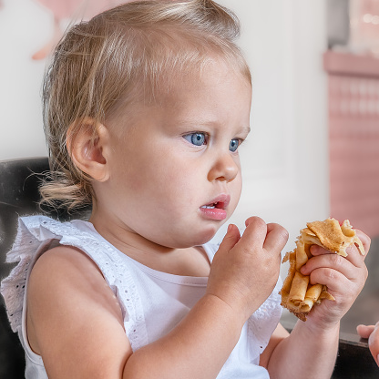 A little girl 1 year old is thoughtfully eating pancakes sitting on a child's chair in a cafe