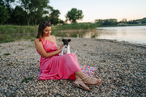 Young woman sitting and having fun with her puppy on lakeshore during summer vacation