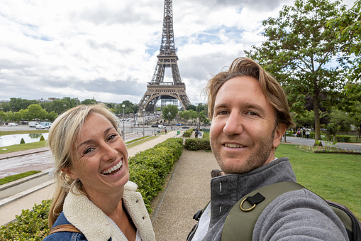 She holds her mobile phone and smiles at the camera, portrait of blond hair woman and man at the international landmark in Paris, France