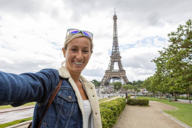 She holds her mobile phone and smiles at the camera, portrait of blond hair woman at the international landmark in Paris, France