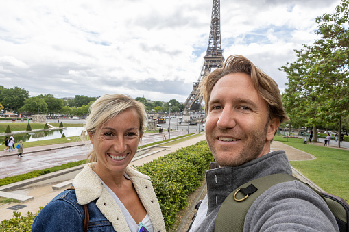 She holds her mobile phone and smiles at the camera, portrait of blond hair woman and man at the international landmark in Paris, France