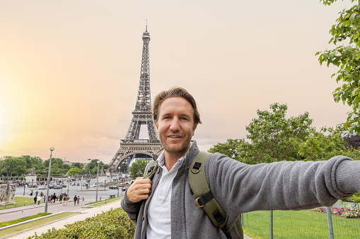 He holds a mobile phone and smiles at the camera, portrait of blond hair man at the international landmark in Paris, France